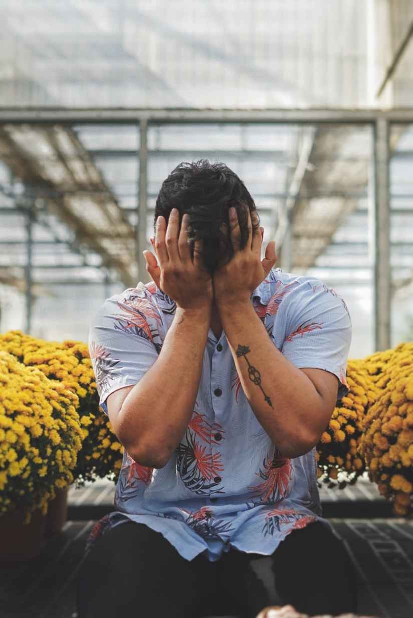 man in floral shirt covering his face with his hands sitting between potted yellow flowers